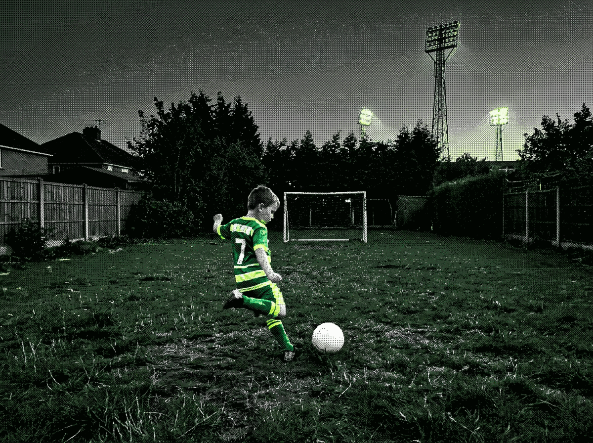 A child in a green kit about to strike a ball in a small floodlit garden, seen through a phone camera